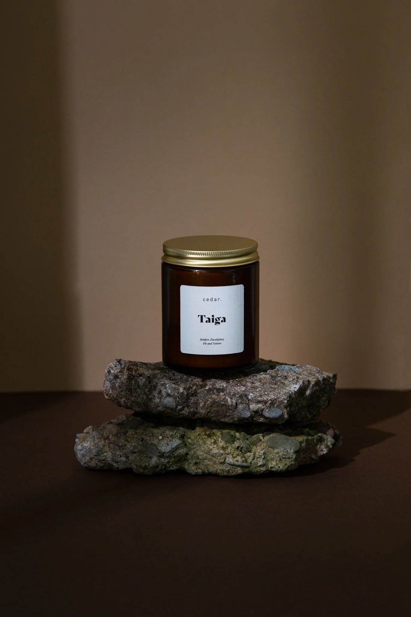 Cedar Lifestyle Taiga Medium Candle in brown glass jar with gold lid standing on 2 pieces of rough rock