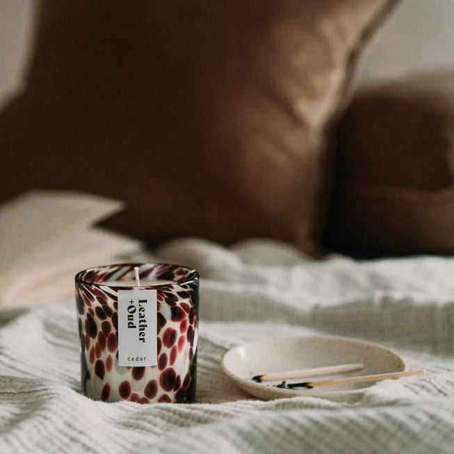 Cedar lifestyles leather and oud scented candle jar on a fabric top next to a dish of burnt matches