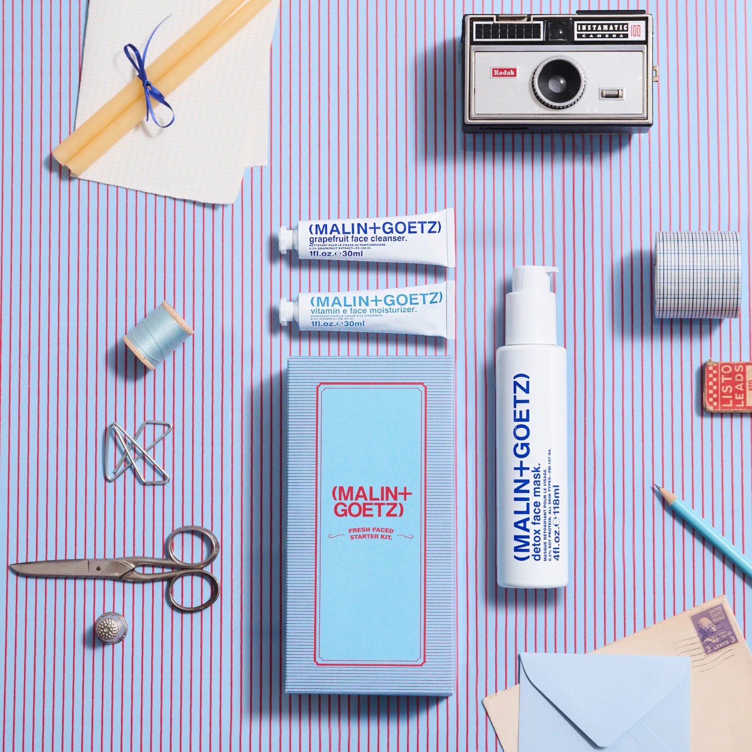 Malin and Goetz Fresh Faced Start Kit 2024 as a flatlay on a blue and red striped background with various stationery items surrounding them
