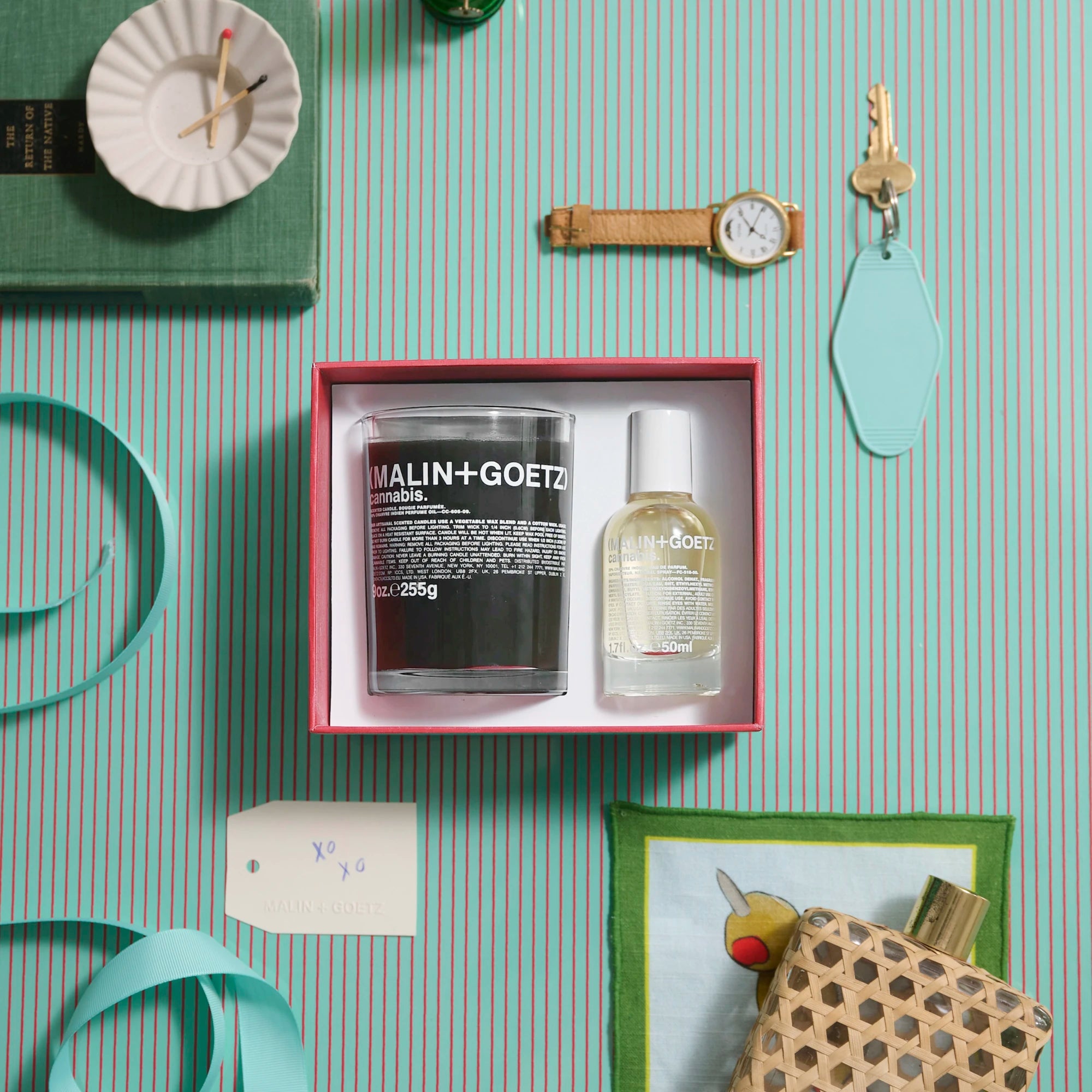 Malin and Goetz Its High Time Cannabis gift set box with candle and perfume bottle. Flat lay image on turquoise and red striped background with personal items laid around it