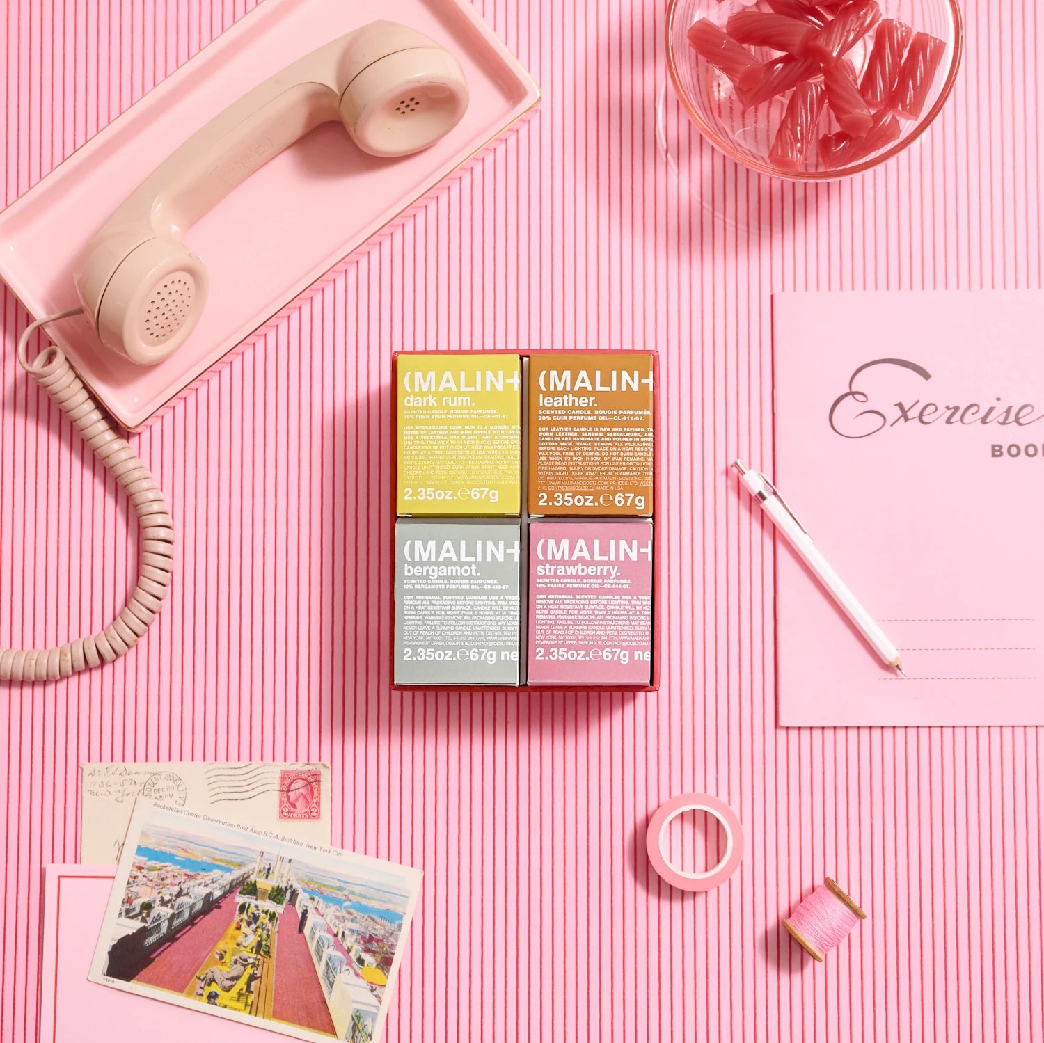 Malin and Goetz Scent The Mood votive gift set as a flat lay image on a pink and red striped background with pink accessories surrounding it