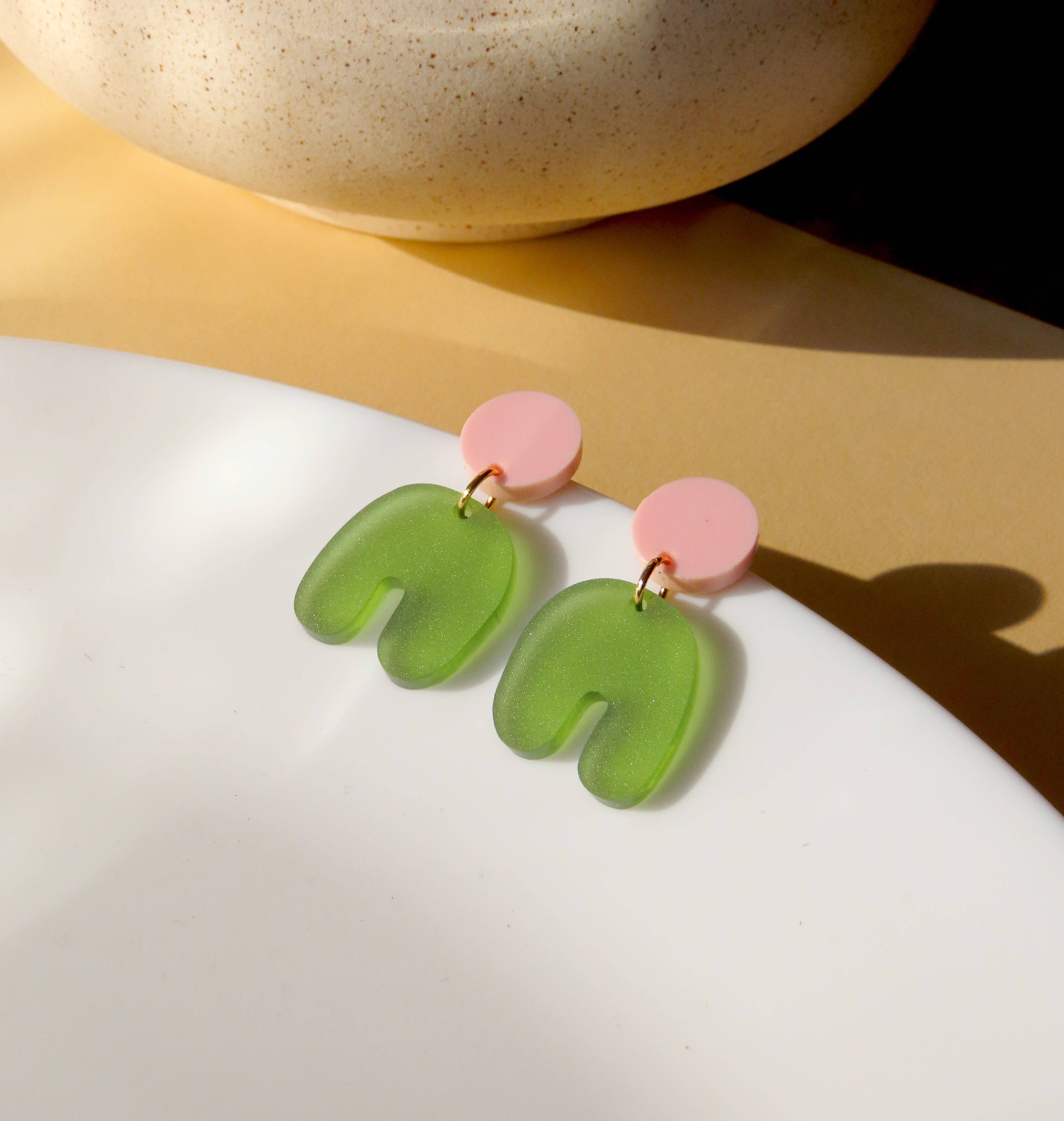 Niemalsmehrohne Squishy Arch Bow Earrings in Pink Olive resting on a white plate with a vase and gold surface in the background