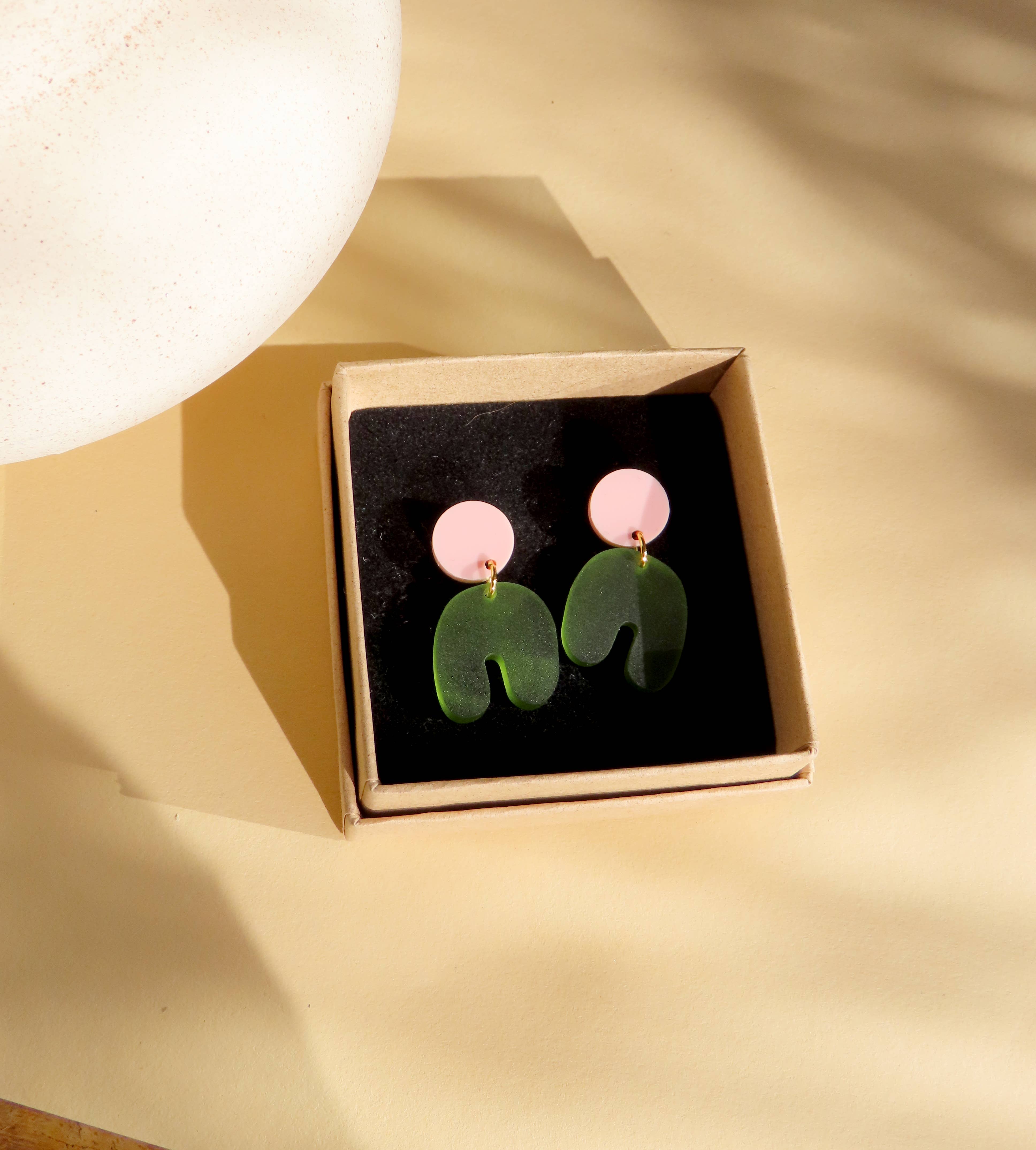 Niemalsmehrohne Squishy Arch Bow Earrings in Pink Olive in their kraft gift box with a gold surface in the background