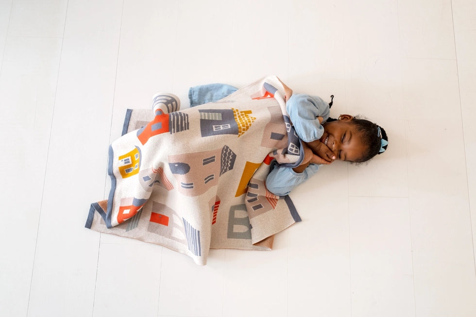 A little girl in a blue dress lying on the floor with the Sophie Home City Cotton Knit Baby Blanket over her