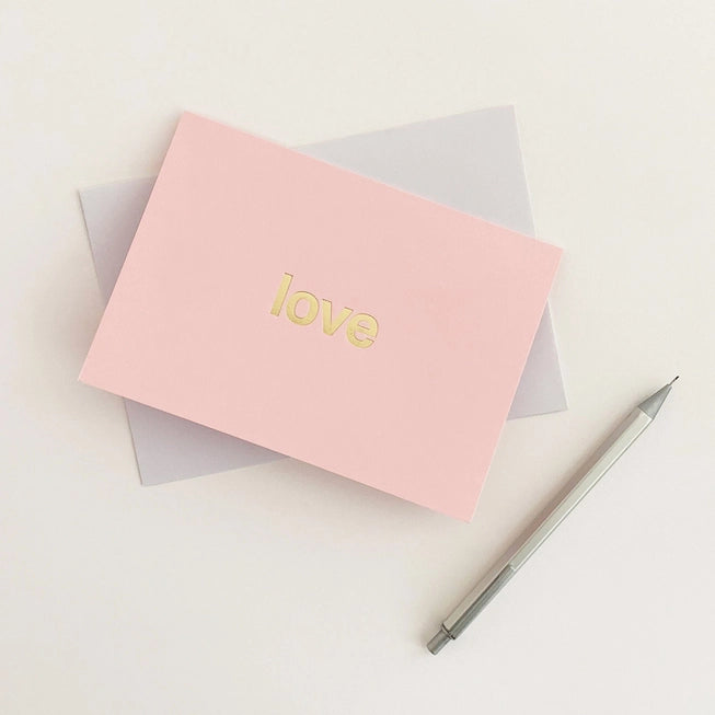 Tom Pigeon brass foiled card with the word love in bold letters on a pink card lying flat next to a silver pen